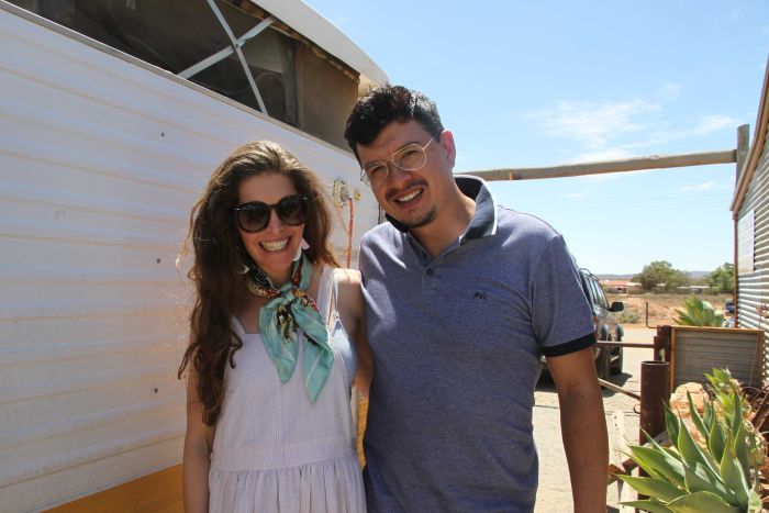 A man and a woman smile at the camera, standing in front of a caravan.